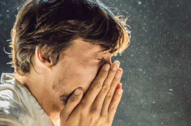 A man sneezing into his hands with visible airborne particles around him, highlighting concerns about indoor air quality for homeowners.