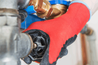 A person wearing a red and black glove turns a valve on a water pipe with a water meter attached, checking for a potential leak—a common concern for homeowners found in many Plumbing FAQs.