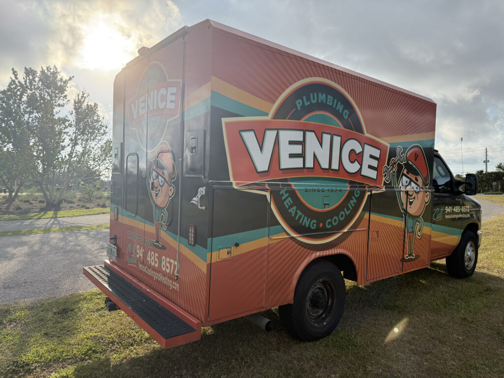 A brightly colored Venice Plumbing Heating Cooling service truck is parked on grass near a road, with trees and sunlight in the background.