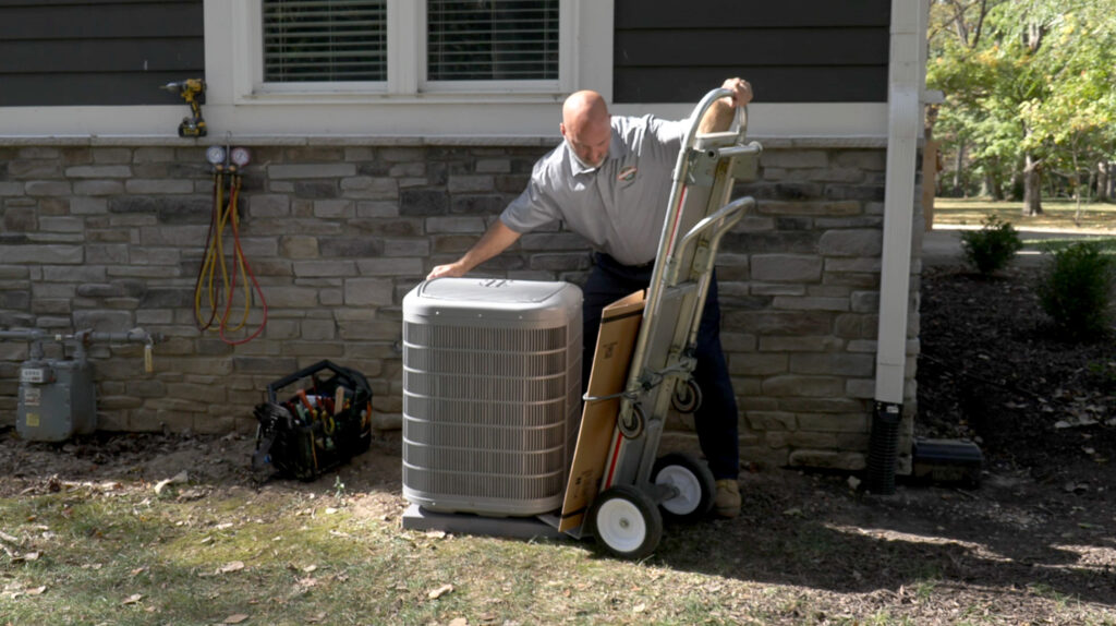 A technician uses a hand truck to move an HVAC unit outside a house, with tools and equipment nearby.