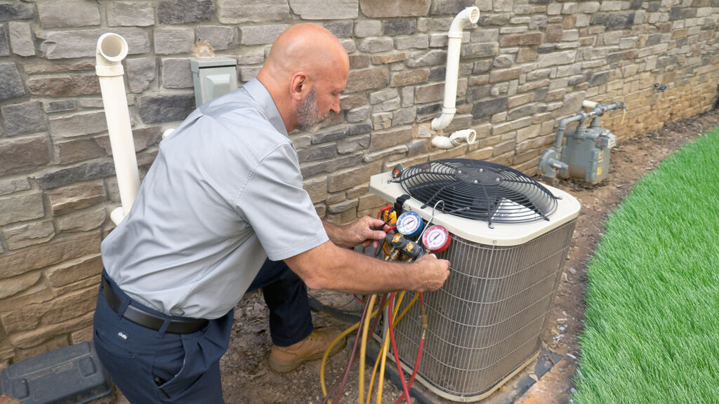 A technician checks the pressure gauges on an outdoor air conditioning unit next to a brick wall.