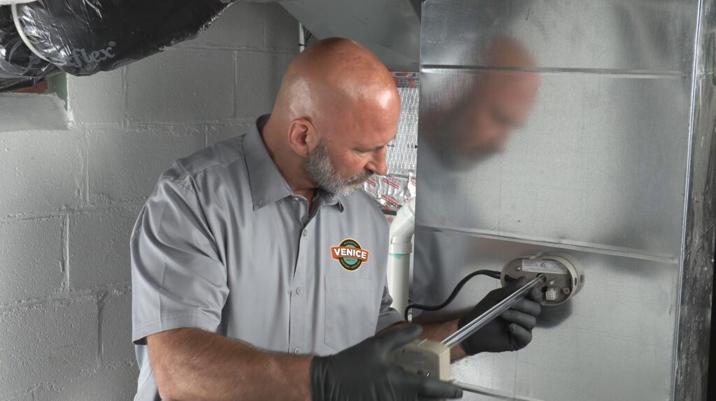 A man in a gray shirt and black gloves installs or services a metal ventilation duct, using a tool to adjust a component attached to the duct.
